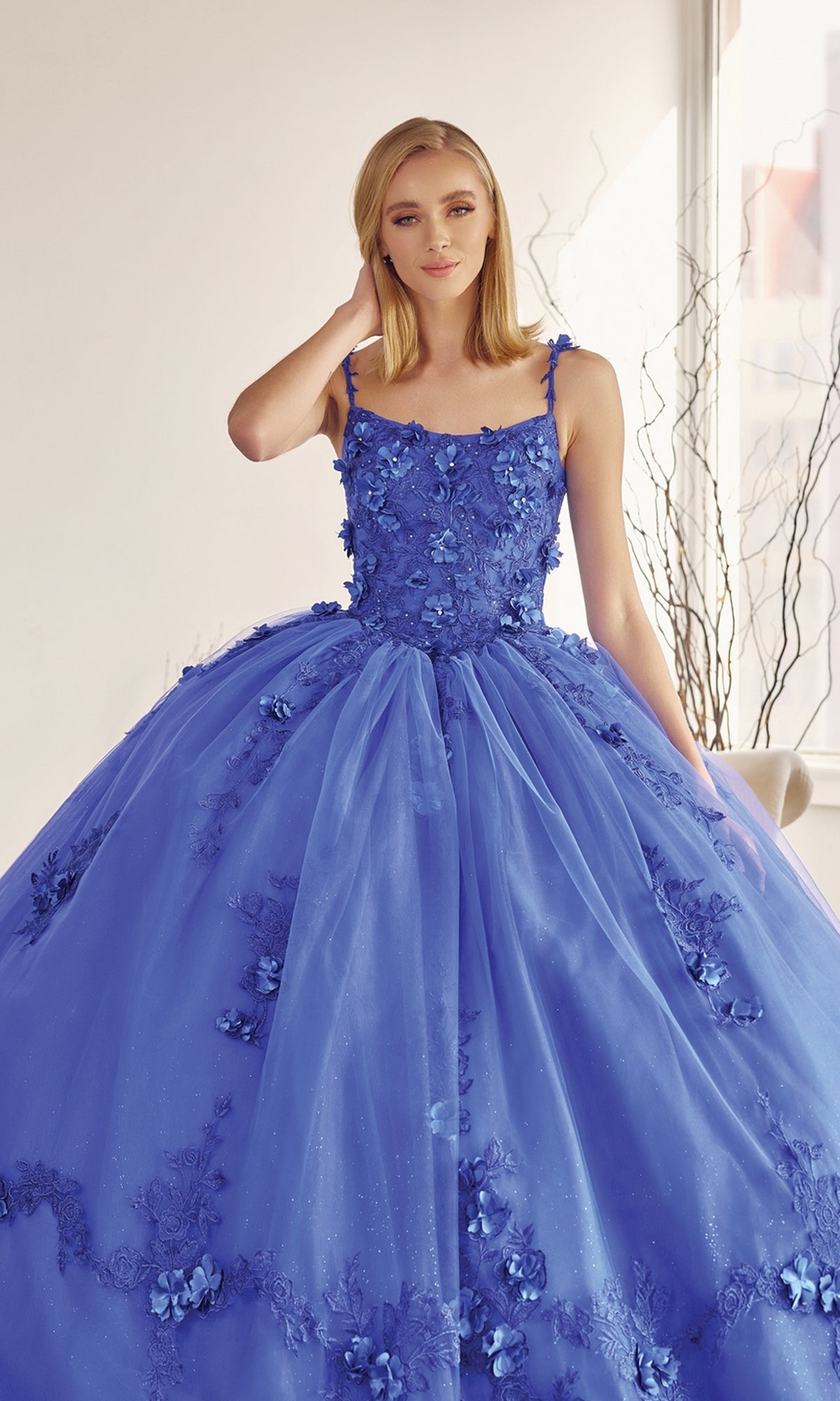 A woman stands indoors wearing the Juliet 1436 Glitter Quinceañera Dress with Cape, featuring floral details and a full tulle skirt.